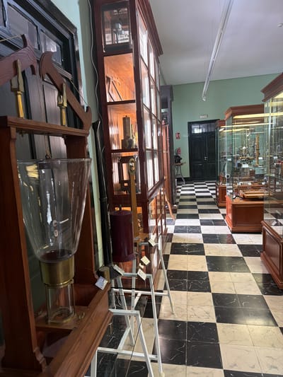 This is a photograph taken in a museum in San Cristóbal De La Laguna, Spain. The scene depicts a display of scientific instruments, likely from a historical collection, housed in wooden display cases with glass fronts. The floor is a striking black and white checkerboard pattern. In the foreground, a large glass bell jar sits within a wooden frame, with visible brass fixtures and what appears to be a pulley system. Behind this, several other display cases are visible, filled with an assortment of antique scientific apparatus, including what look like telescopes, lenses, and other precision instruments. The wall in the background is painted a light green. A dark wooden door is visible in the distance, and a fire extinguisher is mounted on the wall. The lighting appears to be from overhead fluorescent fixtures, typical of a museum environment, suggesting the photograph was taken during operating hours. There is no visible weather or time of day information. The overall impression is one of a well-preserved historical scientific collection, presented in an academic setting. Some small labels with text are visible on the items, but the text is too small to be read.