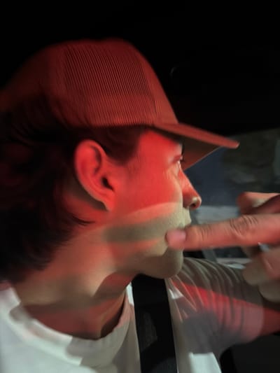 A person wearing a red baseball cap lies on their back. A finger is touching their nose. The person is wearing a white shirt with a black collar. The lighting is dark, with a red tint. The person's face is partially obscured by the shadow of the baseball cap. The background is blurry and indistinct.