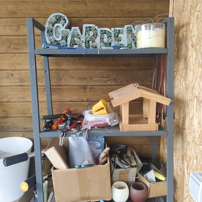 A metal shelf unit is filled with various items, including garden decor, tools, containers, and other miscellaneous objects. On the top shelf, decorative letters spelling "GARDEN" are arranged, filled with artificial succulents. Next to them is a large glass jar containing a yellow substance, possibly a candle. The middle shelf holds a wooden bird feeder, a stack of yellow plastic cups, and a collection of gardening tools such as pliers and secateurs. The bottom shelf contains several cardboard boxes, some open and filled with items, and two decorative pots, one white and one reddish-brown. A white bucket with black handlebars is visible to the left of the shelf. The background consists of wooden plank walls and a wall covered with oriented strand board (OSB). The scene appears to be an indoor storage area, possibly a shed or garage, located in Берега Днп, Russia.