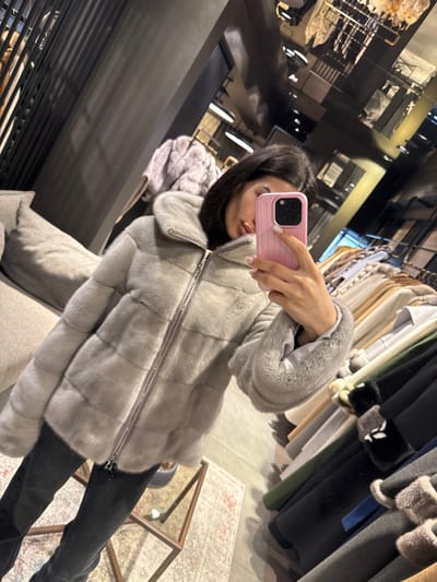 The image displays a mirror selfie of a female subject within a retail establishment in Zeytinburnu, Turkey. The subject wears a light grey, horizontally paneled fur jacket with a central zipper and fur cuffs, paired with dark trousers. She holds a pink smartphone with her right hand, capturing her reflection. The background, viewed through the mirror, reveals an interior consistent with a fur or coat store. Visible elements include a grey upholstered couch, a patterned rug, a glass coffee table, and multiple racks displaying various coats and garments in diverse colors. Overhead, the reflective ceiling shows lighting fixtures and structural components of the room.