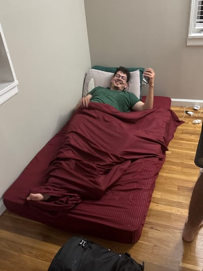 The image captures an indoor scene, likely a bedroom or temporary sleeping area, with a man lying on a bed. The main subject is a young man with dark, curly hair, a beard, and glasses. He is wearing a green t-shirt and is smiling broadly, appearing relaxed and happy. He is lying on a white pillow and is partially covered by a dark red duvet or blanket, which has subtle stripe patterns. He holds a silver-colored smartphone in his right hand, pointing it roughly towards himself, suggesting he might be taking a selfie or engaging in a video call. His bare left foot is visible, poking out from under the blanket at the foot of the bed.
The bed consists of a mattress covered with a dark red fitted sheet, placed directly on a light wooden floor. The room features light grey walls. A window with white blinds is visible in the upper left corner of the frame. In the upper right portion of the image, the bare legs of another person are visible, implying someone else is standing or walking nearby. On the wall near the head of the bed, a cluster of white power outlets with various cords plugged in is visible. A black backpack rests on the wooden floor beside the foot of the bed. The overall impression is candid and informal. No discernible text is visible in the image.