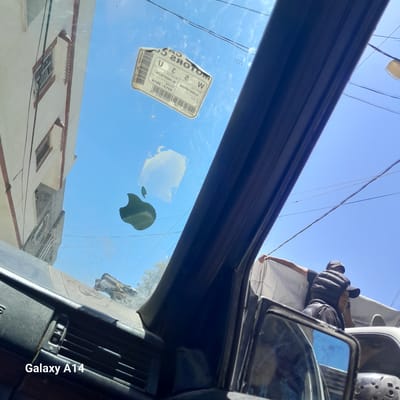 A first-person perspective shot from inside a vehicle looking out through the windshield. The windshield has an Apple logo sticker on it, and a crumpled sticker with Arabic text is adhered to the upper portion. A light-colored building with red trim and barred windows is visible on the left. The sky is clear blue. Outside the vehicle, in the background, a person wearing a black puffer vest over a black hoodie and a baseball cap is standing in the bed of a white truck, holding up a white tarp. Another person is visible behind them. The overall scene suggests an urban environment in Nuseirat, Palestinian Territories, under daylight. The text "Galaxy A14" is overlaid on the bottom left of the image.