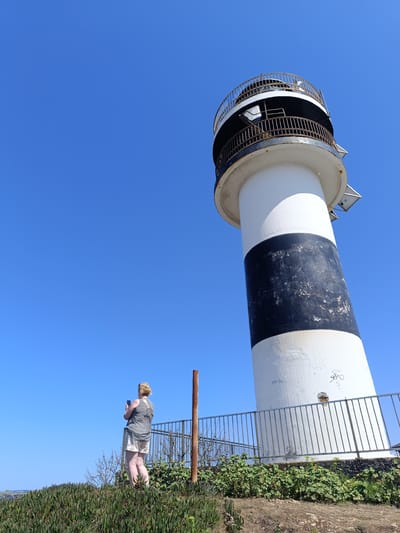 An eye-level, wide shot captures a solitary person standing on a grassy knoll, facing away from the camera and towards a lighthouse. The person, a blonde individual wearing a sleeveless patterned top and light-colored shorts, holds a mobile phone up, seemingly taking a picture or recording a video. The lighthouse, a tall, cylindrical structure with white and black bands, dominates the right side of the frame. Its top section features a circular balcony with a weathered metal railing. The lighthouse stands beside a metal fence that encircles the base of the structure, which is surrounded by lush green vegetation and what appear to be ice plant succulents in the foreground. A rusty metal pole stands vertically to the left of the lighthouse. The scene is set against a clear, vivid blue sky. The image is believed to have been taken in Ribadeo, Spain, at the Faro de Illa Graf.