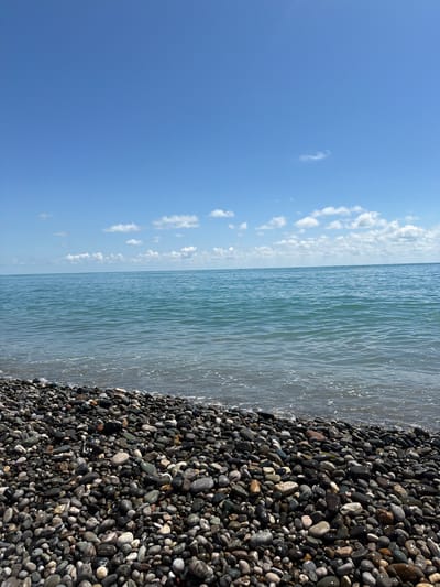 The image depicts a pebble beach on a sunny day in Tsandrypsh, Abkhazia. The foreground is covered with a dense collection of smooth, dark-colored pebbles of varying sizes, glistening from the moisture of the receding waves. The middle ground features the calm, turquoise water of the sea, with gentle ripples and a thin line of white foam at the shore. The horizon is clearly defined, separating the sea from the vast, clear blue sky. Scattered white cumulus clouds drift across the upper portion of the sky. No people or specific objects are visible in the scene, emphasizing the natural landscape.