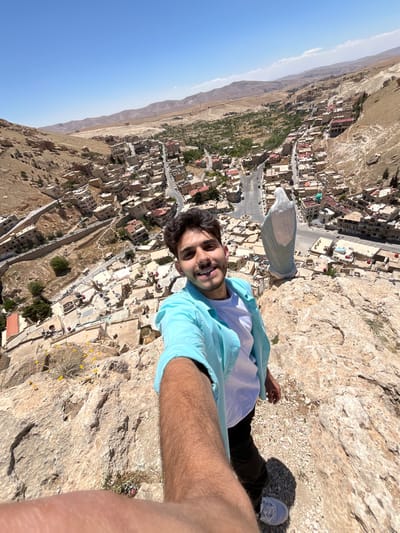 A selfie from a high vantage point depicts a man lying on a rocky outcrop overlooking the ancient city of Ma'loula, Syria. The man has dark hair and a light blue shirt over a white t-shirt. He's positioned so the rocky outcrop appears to be a cliff edge. Below him is a view of Ma'loula, with buildings, roads, and vegetation visible. The sky is clear blue.