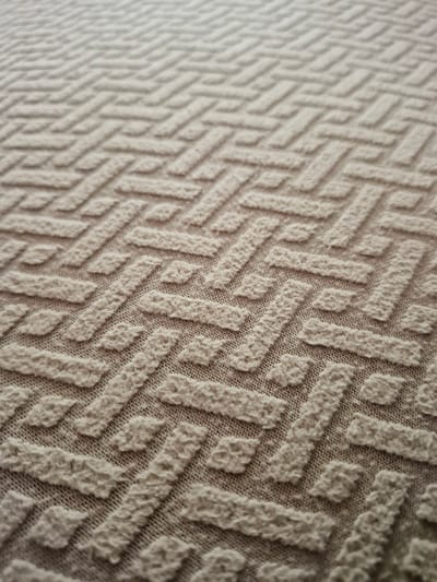 This media file is a close-up, shallow-depth-of-field photograph of a textured fabric. The fabric displays a repeating geometric pattern of raised, interlocking rectangular shapes. The pattern is oriented diagonally across the frame, with the texture becoming less defined and more blurred towards the left side of the image, suggesting a transition from foreground to background or a focus shift. The fabric appears to be a light, neutral color, possibly beige or off-white, with a visible base weave that is darker, creating a contrast with the raised pattern. The texture of the raised elements is rough and irregular, giving the impression of a woven or knitted material with a slightly fuzzy surface. There is no discernible scene, setting, activity, or text present in the image; the focus is entirely on the tactile and visual qualities of the fabric's surface. The lighting appears to be soft and diffused, highlighting the three-dimensionality of the pattern.