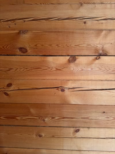 The image displays a close-up, horizontal view of wooden planks. The wood is light brown with visible grain patterns and knots. Some planks show slight cracks or imperfections. The lighting suggests an indoor setting, with some areas appearing brighter than others, possibly due to shadows or light sources not visible in the frame. There are no people, animals, or other objects present in the image. The texture of the wood is rough and natural. The image does not contain any information that would allow for localization to Verkhnevilyuysk, Russia or any specific city.