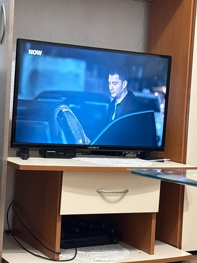 A television displaying a blue-toned image of a man in formal attire is mounted inside a wooden cabinet. The television screen shows the "NOW" logo. The cabinet features an open shelf with an electronic device within. Wires are visible running from the device. The location is Grohotno, Bulgaria.