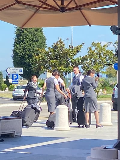 At an outdoor location in Golubovci, Montenegro, five individuals, appearing to be airline crew members based on their attire, are gathered with luggage. The crew members are wearing matching dark grey suits with white shirts and ties or scarves. Two individuals are pulling wheeled suitcases, and another appears to be adjusting her scarf. In the background, a white car is parked, and a directional signpost displays "Čașovno Hourly" and "P" with an arrow. Trees and a bright blue sky form the backdrop. The scene suggests an arrival or departure at an airport or transportation hub.