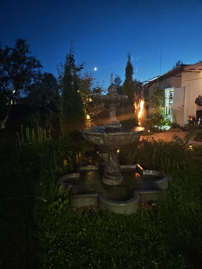 This photograph captures an outdoor scene at dusk, likely in the садоводческое некоммерческое товарищество (SNT) Надежда, Russia. A decorative stone fountain, the central subject, is situated on a patch of green grass. The fountain features multiple tiers, with water flowing into a multi-lobed basin. Beyond the fountain, a building with an open doorway is visible, with a person standing in the background to the right. The sky is a deep blue, indicative of twilight, with a sliver of a crescent moon visible. In the mid-ground, there is a noticeable display of bright orange flames, possibly from a bonfire or pyrotechnics, casting a warm glow on the surrounding foliage and the building. Various trees and tall, slender evergreens are scattered throughout the landscape, and some appear to be adorned with subtle lighting. The overall impression is a garden setting at evening, with an element of fire creating a dynamic visual contrast.