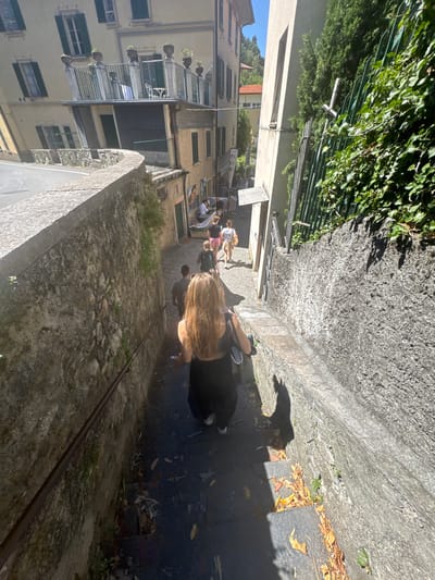 In Gittana, Italy, a woman with long, blonde hair is descending a stone staircase. She is wearing a black tank top and dark pants. Behind her, a man in a dark t-shirt walks up the stairs. Further up the stairs, two women are walking away from the camera; one is wearing a white top and pink shorts, and the other has on a white shirt and a tan skirt. The staircase is flanked by stone walls, with a metal railing on the left side. To the right of the stairs, a green metal fence is overgrown with ivy. The buildings on either side of the staircase appear to be old and made of stone and stucco, with shuttered windows and balconies. The sky is clear and blue, suggesting it is a sunny day. The scene depicts a residential or historic area of the town.