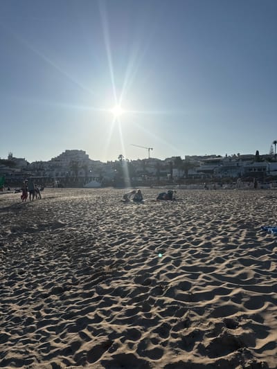 The image depicts a sunny day on a sandy beach in Lagos, Portugal. The sun is shining brightly in the upper center of the frame, casting a strong glare and lens flare. The foreground is dominated by rippled sand dunes, showing texture and shadows. In the midground, several people are visible on the beach. To the left, a small group of people, including children, are walking. Further to the right, two individuals are seated on the sand. In the background, a line of buildings, some with multiple stories and balconies, forms a cityscape against a clear blue sky. Palm trees are interspersed among the buildings, and a construction crane is visible on the left side of the background. Beach umbrellas and loungers are set up in the distance, indicating a resort area. The overall scene conveys a relaxed, holiday atmosphere.