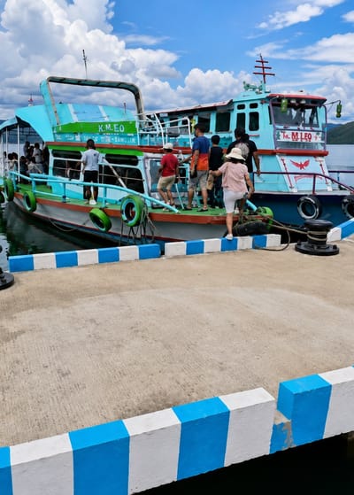 The image depicts a ferry docked at a pier on Lake Toba, specifically at Turpuk Sihotang, Indonesia. The ferry, painted primarily light blue with green and red accents, has "K.M.EO" and "M.LEO" visible on its exterior. Several people are in the process of boarding or disembarking the vessel. On the pier, a woman in a wide-brimmed hat and white shorts is stepping onto the ferry. Behind her, several individuals, including men and children, are also on or near the ramp leading to the boat. The pier is made of concrete and is bordered by a low wall painted with alternating blue and white stripes. The water surrounding the pier is dark, and a distant shoreline with hills is visible under a partly cloudy sky. The overall scene suggests a bustling moment of transit for passengers utilizing this ferry service on the lake.