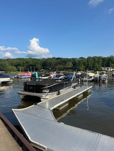 The image shows a view of a marina on a sunny day with a clear blue sky and some white clouds. Several pontoon boats and other watercraft are docked in the water. In the foreground, a silver aluminum dock extends from the left edge of the frame. A black pontoon boat is docked to the right of the dock, with its cover on. In the background, more boats are visible, including some with red and blue hulls, and several jet skis. A line of trees forms the background along the shoreline, with some houses and power lines interspersed. The water is calm, with reflections of the sky and boats visible. The location is the Fox River in Algonquin, Illinois.