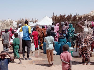 The image depicts a busy outdoor scene in Goshabi, Sudan, centered around a water collection or distribution point. A diverse group of people, including men, women, and numerous children, are gathered and moving through a sandy, dusty environment. The ground shows patches of standing water and mud, suggesting spillage from the containers.

Prominently featured are several large, dark blue plastic barrels and smaller white plastic jerrycans, which are being used for water storage and transport. Many women are dressed in colorful, patterned traditional clothing, often with head coverings, and some are holding small bowls or containers. Men wear various attire, from casual shirts and pants to traditional robes. Children are present throughout the crowd, some observing the activity, while others move about. In the foreground, a child in a dark blue shirt looks towards the viewer, hands near their head, while another child in a red shirt stands with their back to the viewer near the blue barrels.

In the background, temporary structures made of dried straw or reeds are visible, along with a white tent, suggesting a settlement or camp in an arid region. The sky is a clear, light blue, indicating bright daylight, likely midday, given the harsh shadows cast on the ground. Part of a vehicle, possibly a truck bed, can be seen in the mid-left, with the letters "ABU" clearly visible on its side. The scene conveys a communal effort focused on accessing and transporting water.