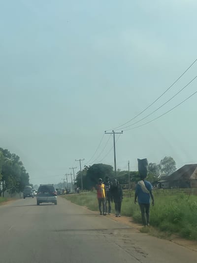 The image depicts a scene on a paved road, likely in the vicinity of Kassa Market, Nigeria, under a hazy, light blue sky. On the right side of the road, a person wearing a blue t-shirt and dark pants walks away from the viewer, carrying a large black bag balanced on their head. To their left, a group of three individuals are walking in the same direction, with one person wearing a yellow and red shirt with the number 55, another in a dark shirt, and a third person whose features are obscured. The road has some visible cracks and imperfections. Several utility poles with power lines run along the side of the road, and lush green grass and vegetation are present. In the background, there are structures that appear to be houses with metal roofs, and trees. Vehicles, including a silver minivan and other cars, are visible on the road ahead. The overall atmosphere suggests an outdoor, daytime scene, possibly during a dry season given the haze.