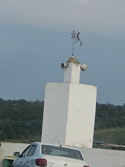 The image depicts a white, square-based minaret situated in the city of Ameur, Morocco. The minaret's upper section features two horn loudspeakers mounted on opposite sides and is topped with a conical cap from which a metallic finial, shaped as a crescent moon and a five-pointed star, protrudes. The minaret shows signs of weathering, including dark streaks and discoloration on its white surface. In the foreground, partially visible below the minaret and to the left, is the rear section of a white Dacia sedan, identifiable by its taillights and the visible "Dacia" emblem and "V" series designation on the trunk lid. The vehicle appears to be positioned near a low, white concrete barrier or wall. The background consists of rolling hills densely covered in green vegetation, indicative of a forested or vegetated landscape, under an overcast, light grey-blue sky. No individuals are present or engaged in any discernible actions or interactions within the frame.