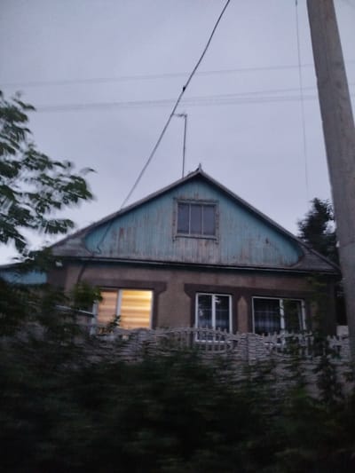 The image shows a two-story house with a blue, weathered wooden gable. A window is visible in the gable, and two windows are on the floor below. One of the lower windows is illuminated from within, suggesting it is evening or night. The house is partially obscured by dark green foliage in the foreground. A concrete utility pole and wires are visible to the right of the house. The sky is overcast, indicating a cloudy day or twilight. The overall scene suggests a rural or suburban setting, likely in Ak-Suu, Kyrgyzstan, given the provided location context. There is no visible text in the image.
