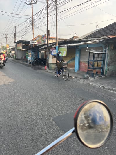 A street scene is depicted in Sugih Waras, Indonesia, characterized by a paved road with patchy grey asphalt, bordered by a row of small shops and houses. The sky is overcast with a hazy, light grey tone, suggesting either early morning or late afternoon. Overhead, a dense network of electrical wires is prominently displayed, supported by wooden poles.
In the foreground, the reflection of a vehicle, likely a motorcycle given the perspective from its side mirror, is visible. The mirror, with a red rim, shows a blurry reflection of the road, buildings, and a darker vehicle.
Further down the street, a woman wearing a green hijab and a face mask is riding a bicycle. She is dressed in a long, dark green tunic and pants, with blue sandals on her feet. The bicycle is dark blue and has a basket in the front.
To the left of the woman, a few motorcycles are parked. The shops lining the street appear to be open, with various signs and advertisements visible. One sign reads "TOKO AKI", indicating a battery shop. Another sign, partially obscured, appears to advertise a "RE-TECH PERTAMA!" service. The buildings have corrugated metal roofs and some feature blue painted facades.
On the right side of the street, a wire cage structure, possibly for animals or birds, stands near the sidewalk. There are also signs of litter on the ground. The overall environment suggests a typical urban or semi-urban commercial street in Indonesia.