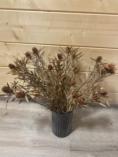 A dried floral arrangement featuring brown, cone-shaped blooms and narrow leaves is displayed in a dark, ribbed vase. The vase sits on a light-colored, wood-plank floor. Behind the arrangement is a wall constructed of light-colored wood planks, horizontally oriented. The scene appears to be indoors, possibly in a home or studio. The lighting is soft, casting a subtle shadow from the vase onto the floor. There are no people or animals present in the image. The location is not specified in the image itself, and it is therefore not possible to determine if it is in the city of Карабаново, Russia.
