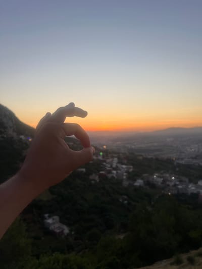 The image, correctly viewed horizontally, captures a twilight or sunset scene overlooking the city of Tétouan, Morocco, from a high vantage point. In the foreground, a hand is prominently displayed, forming a gesture where the thumb and index finger touch to create a circle, often associated with a peaceful or "OK" sign. The hand is partially silhouetted against the bright horizon, indicating the light source is behind it.

The sky presents a striking gradient of colors, characteristic of sunset or twilight. A warm band of orange, yellow, and soft red hues dominates the horizon line, transitioning upwards into a clear, lighter blue-grey sky. Below the horizon, the sprawling urban landscape of Tétouan is visible, extending into the distance. Buildings and city lights, appearing as blurred bokeh points, dot the scene, especially noticeable against the darkening terrain. Dark, tree-covered hills or mountains frame the lower and right parts of the image, separating the city from the viewer's immediate position. The overall atmosphere is one of tranquility and contemplation, emphasizing the beauty of the dusk hour over the Moroccan landscape. No visible text is present.