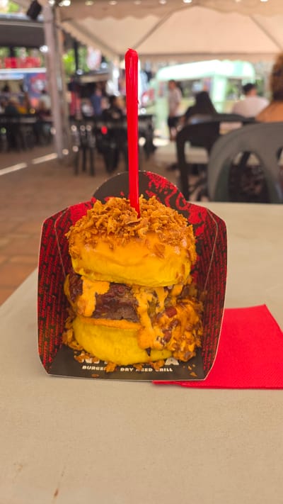 This is a close-up, slightly tilted, eye-level, outdoor shot of a burger served in a black and red paper container, possibly from a food stall or casual eatery, with the text "BURGER DRY AGED GRILL" visible on the container's edge. The burger is held together by a red plastic skewer and is topped with what appear to be crispy fried onions and a generous drizzle of orange sauce, possibly cheese sauce or a spicy aioli. The bun is golden yellow and appears soft.
In the background, the scene is blurred, suggesting an outdoor setting, likely a public space like a street market or a plaza, given the presence of other people and what looks like white tenting or awnings, common in outdoor events or temporary food setups. The lighting suggests it's daytime, possibly late morning or afternoon, with diffused light. The overall ambiance is casual and focused on the food. The vibrant red of the skewer and the napkin peeking out from the right add pops of color to the image. The visible text on the packaging indicates the burger is from a place that specializes in dry-aged burgers.