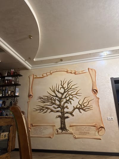This is a photograph of a wall mural depicting a tree with scrolled documents. The mural is rendered in earthy tones, with a stylized tree branching across a beige background that resembles parchment. Two rolled scrolls are integrated into the mural; one on the left and one on the right.
The scroll on the left has a series of squares cascading down it, with numbers "80" and "16" visible on two of them. The scroll on the right contains text written in Armenian script. Below the mural, a dark wooden structure with shelves holds several bottles, likely wine or spirits, suggesting the mural is located in a residential or commercial establishment like a restaurant or cellar.
The wall itself has a textured finish, and the lighting suggests an indoor setting, possibly illuminated by artificial lights. There's a ceiling with decorative molding and crystal accents visible in the upper left portion of the image, indicating a well-decorated interior. The overall mood is one of history and lineage, with the tree symbolizing family or a lineage and the scrolls perhaps representing historical records or family history. The Armenian text is a notable detail, hinting at a specific cultural context. There are no people visible in the image, only the mural and the surrounding architectural elements.