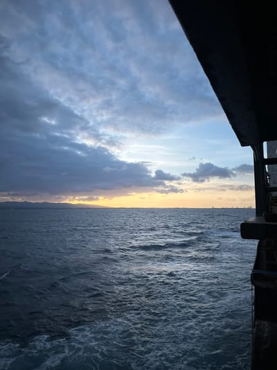 This image depicts a vast expanse of the ocean under a cloudy sky, likely taken from the deck of a vessel. The water is a deep blue, with visible waves and white foam churning on the surface. On the horizon, a faint line of land with mountains is visible, bathed in the warm glow of a sunset or sunrise. The sky is filled with dramatic clouds, varying in color from dark grays to lighter blues and hints of orange and yellow from the setting or rising sun. To the right, the dark silhouette of the vessel's structure intrudes into the frame, suggesting a perspective from a ship or ferry. There are no people visible in the scene, and the focus is on the natural elements of the sea and sky, as well as the imposing structure of the vessel. The overall atmosphere is serene yet dramatic, with the interplay of light and shadow across the clouds and water. The location in the city of Le Gosier, Guadeloupe, is not discernible from the visual elements present in the image.