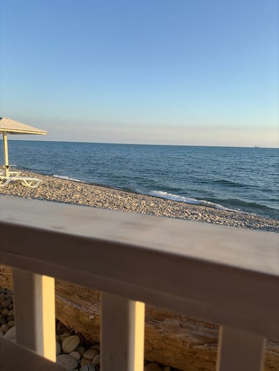 This is a low-angle, landscape-oriented photograph taken at eye-level, capturing a serene beach scene in Shirokaya Balka, Russia. The image is oriented vertically, with the horizon line running diagonally from the bottom left to the top right.

The foreground is dominated by a light-colored wooden railing, which is partially out of focus and occupies the right side of the frame. Beyond the railing, the scene unfolds to reveal a pebbled beach. The pebbles are smooth and vary in shades of gray, white, and beige, some appearing wet from the receding tide.

The middle ground features the calm, blue-gray water of the sea, with gentle waves lapping at the shore, creating a thin line of white foam. The water's surface is textured with ripples. In the far distance, a faint outline of what appears to be a boat or a distant structure can be seen on the horizon.

The sky occupies the upper left portion of the image and transitions from a clear, light blue at the very top to a softer, pale yellow or white hue near the horizon. The overall lighting suggests it is either early morning or late afternoon, casting a warm, soft glow on the scene. The weather appears calm and pleasant, with no discernible clouds.

The image conveys a sense of tranquility and natural beauty. There are no visible people or specific events taking place, allowing the viewer to focus on the peaceful coexistence of the sea, shore, and man-made railing. No text is visible in the image.