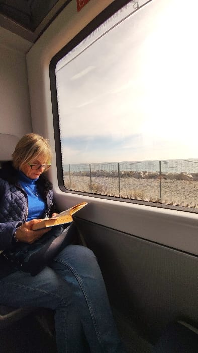 An adult female is seated within a train carriage, reading an open book with a light-colored cover. She wears glasses, a blue quilted jacket with a dark fur collar, a blue turtleneck, and blue jeans. Her blonde hair is styled short. The train's interior features a dark grey window frame and light grey paneling. Outside the window, a bright, hazy sky hangs over a body of water, likely the Mediterranean Sea, adjacent to Cabrera de Mar, Spain. A sandy/rocky shoreline is visible between the train and the sea, bordered by a metal fence composed of vertical posts and horizontal mesh. Several large rocks are present on the beach side of the fence.