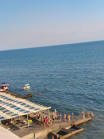 A clear, bright day at a beach resort on the coast of Blagoveschenskaya, Russia, with people enjoying the water and the sun. The scene is set along a concrete pier and a stretch of beach with many lounge chairs under white and blue striped awnings. The vast, calm blue sea fills the majority of the frame, meeting a clear, pale blue sky at the horizon.

On the water, a few small boats are visible, including a pedal boat with several people aboard, and two small red and white recreational boats docked near the pier. Scattered throughout the water are numerous people swimming, some appearing to be children.

Along the pier, a diverse group of people are gathered. Some are standing and chatting, while others are sunbathing on lounge chairs. A few are entering or exiting the water. One man in a white shirt and dark shorts stands near a life preserver. Children can be seen playing. A bright yellow inflatable ring rests on the pier.

The time of day appears to be late afternoon, given the angle of the sunlight casting shadows. The weather is fair and sunny. The overall mood is relaxed and recreational, with people enjoying a summer day at the seaside. There is no visible text in the image.