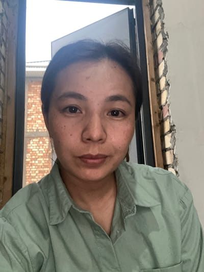A medium shot photograph shows a woman with dark hair, fair skin with freckles, and dark eyes, looking directly at the camera. She is wearing a light green collared shirt. The background is out of focus, showing a brick wall and window frame. Based on the provided metadata, the image was taken in Novopokrovka, Kyrgyzstan.