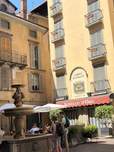 The image depicts a quaint courtyard scene in the city of Mozzo, Italy, bathed in bright sunlight. The architecture features weathered yellow stucco buildings with multiple stories, adorned with balconies featuring intricate wrought-iron railings. Several windows are visible, many with closed wooden shutters in a muted blue-gray color. A prominent feature is an antique stone fountain in the foreground, its tiered basin exhibiting intricate carvings.
In the background, an establishment named "Albergo Ristorante Agnello D'Oro" is identifiable by a sign above an arched entrance. A red awning with the same name extends over the entrance to the restaurant. The area appears to be a public space with several tables and umbrellas set up, suggesting an outdoor dining area.
Two individuals are standing in the foreground, facing away from the camera, appearing to be tourists. One person wears a light green t-shirt and shorts, carrying a blue backpack. The other wears a dark top and shorts, also with a backpack. Several other people are seated at tables in the background, some engaged in conversation, others looking at their phones. Greenery in planters adds a touch of life to the scene. The overall atmosphere is one of a relaxed European town square.