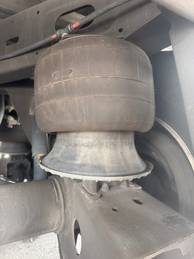 An air spring for a vehicle's suspension system is shown mounted between the frame and axle. The air spring is a black, cylindrical bellows that has text visible on its surface, which appears to read "Firestone." A thin black hose with a red wire inside is connected to the top of the air spring. The air spring is attached to a metal bracket on the vehicle's frame, and a rubber boot extends from the bottom of the air spring to a metal component of the axle. The vehicle is parked outdoors, likely on an asphalt surface, with a blurred background that suggests an urban or suburban environment. No people or specific landmarks are identifiable, and the image does not provide enough information to pinpoint the location to the city of Lebanon, United States of America.