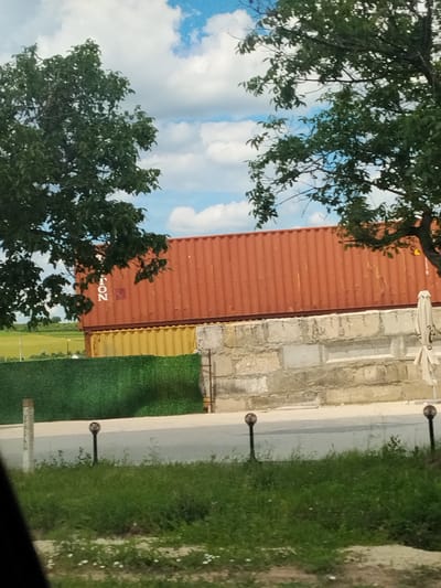 The image depicts an outdoor scene, possibly in Budești, Moldova, featuring stacked shipping containers and natural elements. In the foreground, a grassy area with small white flowers is visible, bordering a paved surface. Two small, dark-colored post lights with spherical tops are positioned on either side of the paved area, suggesting a pathway or road. Behind the grassy area and paved surface, a vibrant green, textured fence is present, extending horizontally across the frame.
Behind the green fence, two shipping containers are stacked. The bottom container is a muted yellow, and the top container is a deep orange-red. The orange-red container has visible corrugated siding and some markings, including the letters "T", "O", "N" arranged vertically, and a partial red square logo.
Further in the background, a wall constructed of rough, rectangular concrete blocks is visible, partially obscuring the lower portion of the yellow shipping container. A light-colored, folded umbrella or canopy structure is positioned to the right of this wall. Beyond the containers and wall, a flat, green field stretches towards the horizon under a partly cloudy blue sky. Two trees with dark green foliage frame the top portion of the image, with their branches extending over the shipping containers and the sky. The overall impression is one of an industrial or storage area juxtaposed with nature.