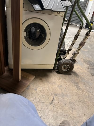 The image shows a white washing machine being moved on a green hand truck. The washing machine is partially obscured by a wooden furniture piece on the left. The hand truck has two wheels in the front and two smaller wheels at the rear. The handles of the hand truck are wrapped in aged tape. The scene appears to be indoors, possibly in a garage or storage area, with a concrete floor and a grey metal garage door in the background. The lighting suggests it is daytime. No people are visible in the frame. The overall impression is of an appliance being relocated or stored. The image does not contain any discernible landmarks or details that would allow for precise location identification within Osnabrück, Germany.