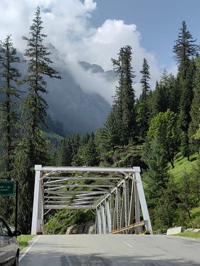 The image depicts a road leading to a white steel truss bridge in Kothi, India. Tall evergreen trees line the road and flank the bridge, with a steep, verdant mountainside visible in the background. The mountains are partially obscured by clouds and mist, creating a dramatic, atmospheric scene. A green sign with white lettering is mounted on a post to the left of the road, indicating "Kothi-2 Bridge". A grey car is parked on the left side of the road, partially visible in the foreground. The sky is partly cloudy with patches of blue visible.