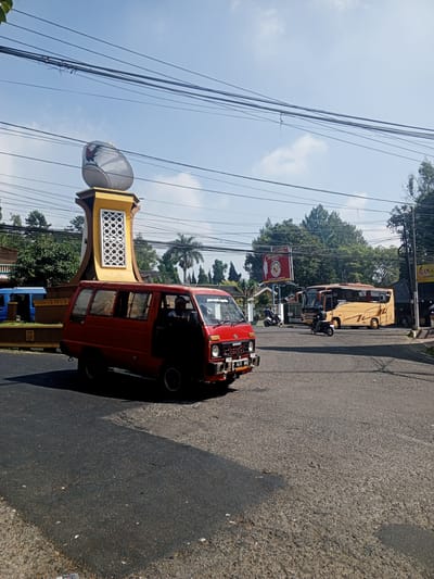 The image depicts a street scene in Ruwan Eliya, Sri Lanka, under a clear blue sky with scattered clouds. A prominent monument, featuring a large, egg-shaped object with a rooster emblem at the top, stands on a yellow and brown pedestal. The monument is adorned with intricate white geometric patterns.

In the foreground, a red van is parked on the asphalt road. The van has its doors slightly ajar and a person is visible in the driver's seat. The license plate of the van is partially visible and reads "F 1417 HA".

Further down the road, a large yellow and brown bus is seen. Two individuals on motorcycles are also present in the scene, one of whom appears to be riding a scooter. In the background, several trees and buildings are visible, including a blue van parked near the monument. Overhead, a network of power lines crisscrosses the sky. The overall environment suggests a sunny day in a public area with vehicular traffic and urban elements.