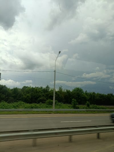 This is a photograph taken from a moving vehicle, looking out at a roadside scene. The sky is overcast with heavy, grey clouds. In the middle ground, there is a dense line of green trees. Beyond the trees, there appear to be power lines and a lamppost. In the foreground, a highway is visible with a yellow line dividing the lanes. A silver guardrail runs along the edge of the road. A dark-colored vehicle is visible in the far right lane of the highway, blurred due to motion. The location is identified as Molkino, Russia.