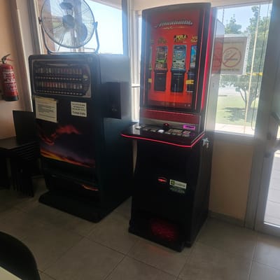 The image depicts an indoor scene in Barrio Estación, Spain, featuring two large machines. On the left is a dark-colored vending machine with a display showing rows of cigarette packs. A fire extinguisher is mounted on the wall to the left of the vending machine. To the right is a red and black gaming machine with a screen displaying various slot machine games. The gaming machine has a control panel with buttons and a coin slot. Both machines are positioned near a large window that looks out onto a grassy area with trees. A ceiling fan is visible above the vending machine. Several signs are attached to the machines and the window, some of which are in Spanish and relate to health warnings and regulations. The floor is tiled. There are no people visible in the image.