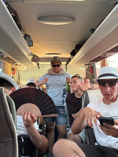 The image depicts a group of men inside a bus, likely a tour bus or public transport. The men are in their early twenties and are wearing casual attire, including t-shirts, shorts, sunglasses, and hats. One of the men is holding a bottle of beer, while another is holding a fan. It is not possible to determine if the city of Cavallino, Italy is definitively visible in this photo.