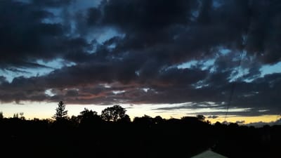 The image depicts a twilight sky with dramatic clouds, as seen from Romny, Ukraine. The sun is setting, casting a warm, yellow glow near the horizon, which is obscured by a silhouetted tree line and what appears to be a structure with a sloping roof. Dark, heavy clouds fill most of the frame, interspersed with patches of lighter, illuminated clouds that show hints of pink and orange from the fading sunlight. The lower portion of the sky transitions into deep blues and grays, suggesting the approach of night. Two thin, dark lines, likely power lines, run vertically through the left side of the image, creating a sense of depth and context. The overall mood is atmospheric and somewhat somber, characteristic of a cloudy sunset. There is no visible text in the image.