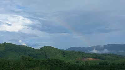 The image shows a landscape in Ban Tak Daet, Thailand, with green, forested hills in the foreground and middle ground. In the background, taller hills are partially obscured by clouds. A faint rainbow arches across the sky to the right of the center. The sky is filled with clouds. Some cleared land is visible at the base of one of the hills.