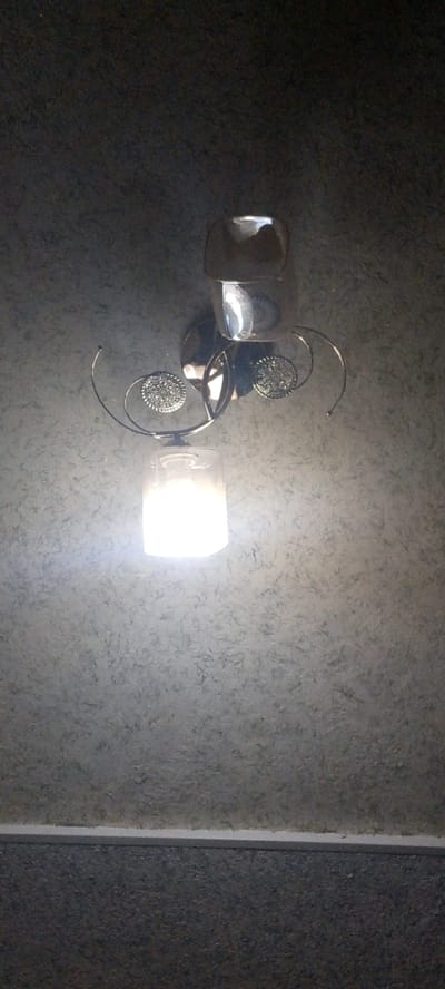The image shows a close-up view of a wall-mounted light fixture. The fixture is made of metal with a shiny, chrome-like finish and features decorative elements resembling ornate circular medallions. A glass shade, currently illuminated, hangs below the main body of the fixture. The wall behind the fixture has a textured, mottled gray finish. The lower portion of the image reveals a horizontal white trim separating the textured wall from a darker, rougher surface below. The scene appears to be indoors. The location is identified as Marhamat, Uzbekistan.
