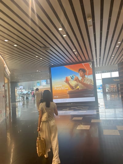 The image captures an interior view of a modern, well-lit airport terminal in Guangzhou City, China. The ceiling features a distinctive slatted design with integrated lighting. In the foreground, a woman is walking away from the camera, carrying two shopping bags. She is wearing a light-colored outfit, including a t-shirt and loose-fitting pants. To her right, a large digital billboard displays an advertisement for the HUAWEI Pura 9 series, featuring a young man holding the phone. Further down the terminal, other travelers are visible, some with luggage, walking in the same direction. Shops are lining the sides of the walkway, with one clearly labeled "Vivien." The floor is highly reflective, with a checkered pattern of dark and light tiles, reflecting the overhead lights and the surrounding environment. In the background, through large windows, a portion of an airplane is visible, suggesting the proximity to the tarmac. The overall atmosphere is one of a busy, contemporary transportation hub.