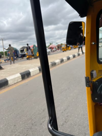 This is a first-person view from a yellow auto-rickshaw, likely in Gulum, Nigeria. The scene depicts a street with a marketplace on the left side. Several people are visible, some appearing to be vendors and shoppers. Auto-rickshaws and other vehicles are present in the background, and market stalls with umbrellas are set up. The sky is overcast with clouds. The auto-rickshaw is in motion, indicated by the blurred road and the perspective. The focus is on the immediate surroundings of the auto-rickshaw, with a black metal bar dominating the foreground on the left. The road has a yellow line marking and a black and white striped curb separating it from the pedestrian area.