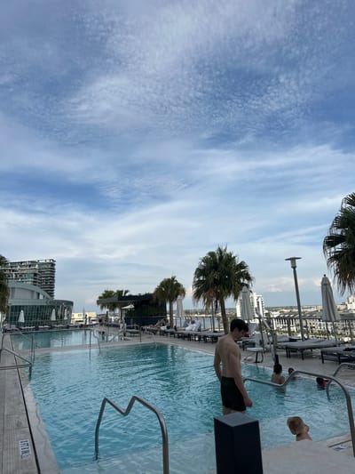 The image captures an outdoor swimming pool scene in Tampa, Florida. The pool water appears clear and blue, reflecting the sky. Several individuals are present, including a man standing near the edge of the pool with his back to the camera, wearing dark shorts. Other people are swimming in the pool and sunbathing on lounge chairs nearby. Palm trees provide some shade along the pool deck. The pool is part of a high-rise building. The sky shows broken clouds with a blue hue. The pool has metal handrails and a concrete deck. In the background, other city buildings are visible.