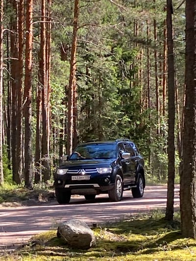 A black Mitsubishi Pajero Sport SUV is driving on a dirt road in a pine forest. The vehicle is facing forward and is partially obscured by trees. The sun is shining, casting shadows on the road and the surrounding mossy ground. A large rock is visible in the foreground. The license plate of the vehicle, Y433HY 178, is partially visible. The image appears to have been taken in the city of Leipyasuo, Russia.