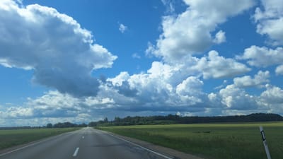 A paved, two-lane road stretches into the distance under a bright blue sky filled with large, white cumulus clouds. The road has solid white lines on the edges and dashed white lines down the center. On the left side of the road is a green field with some scattered trees in the distance. On the right side of the road is a wider green field bordered by a dense forest. A white signpost with black numbers "1" and "9" is visible in the foreground on the right. The scene appears to be a rural landscape in Ogresgals, Latvia.