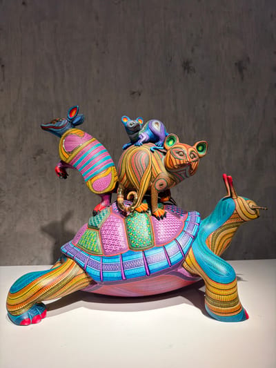 This media file captures a vibrant, intricately carved and painted alebrije sculpture, a traditional form of Mexican folk art, originating from San Martín Tilcajete, Mexico. The sculpture features multiple fantastical animals stacked and interconnected.
The central and largest component is a multi-colored turtle, serving as a base. Its shell is adorned with geometric patterns and swirls in shades of purple, blue, green, and pink. Perched atop the turtle is a larger animal, possibly a stylized fox or feline, with prominent green-rimmed eyes. Its body is covered in flowing yellow and orange patterns. Clinging to its back are two smaller, similarly stylized creatures, one purple and one blue, both with large eyes and detailed patterns. Extending from the lower-left side of the turtle is an armadillo-like animal, characterized by pink and blue stripes and a patterned body. Another creature, resembling a bird with a long beak and blue and yellow plumage, extends from the upper-right of the turtle.
All parts of the sculpture are meticulously decorated with fine lines, dots, spirals, and geometric motifs in a dazzling array of bright, contrasting colors. The polished finish of the wood (or similar material) enhances the vividness of the paintwork.
The sculpture is displayed indoors, positioned vertically against a corner formed by two walls. The wall on the right is a plain, light grey, while the wall on the left is darker with a subtle texture, suggesting a gallery or exhibition space. Artificial lighting illuminates the piece, casting shadows that emphasize its three-dimensional form. No people are visible, and there is no visible text. The image focuses entirely on the elaborate artistry of the alebrije.